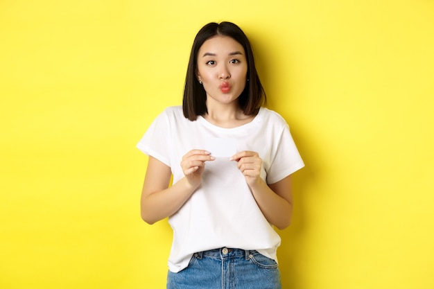 Junge asiatische Frau in lässigem weißem T-Shirt mit Plastikkreditkarte und albernen Lippen, beim Einkaufen, gelber Hintergrund