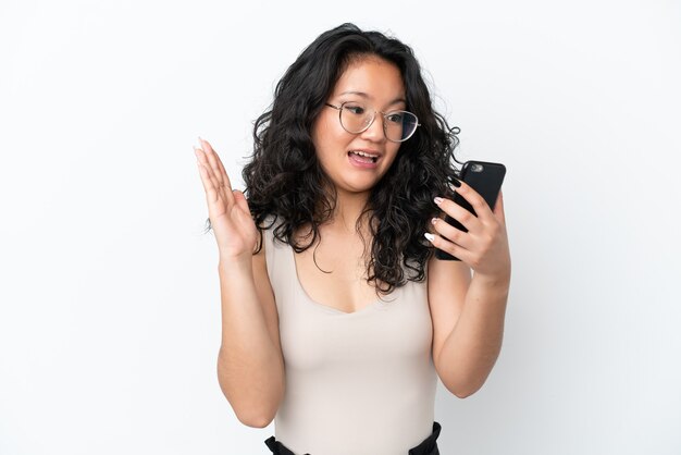 Junge asiatische Frau isoliert auf weißem Hintergrund in die Kamera schaut, während sie das Handy mit überraschtem Ausdruck benutzt