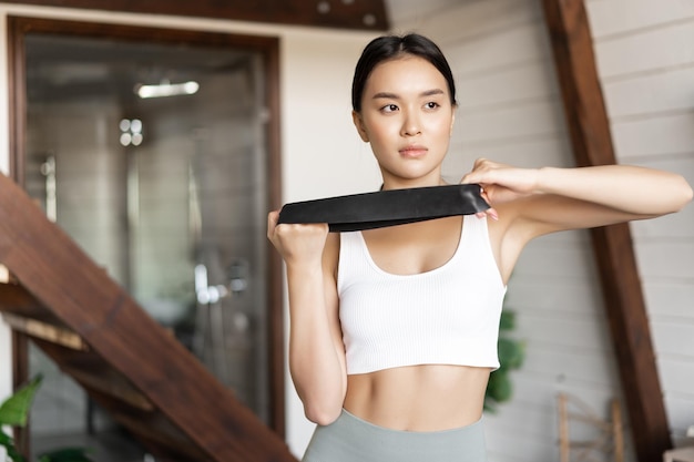 Junge asiatische frau macht zu hause fitnesstraining mit elastischem seil-dehnband für arm-bizeps-sta...