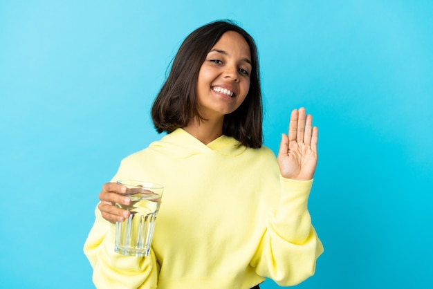Junge asiatische Frau mit einem Glas Wasser lokalisiert auf blauem Hintergrund, der mit Hand mit glücklichem Ausdruck salutiert