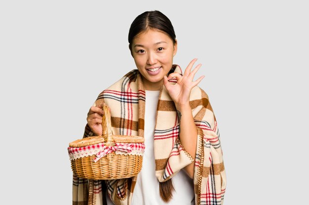 Junge asiatische Frau mit einem Picknickkorb, isolierte, fröhliche und selbstbewusste, zeigt eine ok-Geste