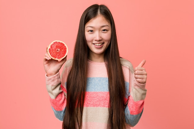 Junge asiatische Frau mit einer Pampelmuse oben lächelnd und Daumen anhebend