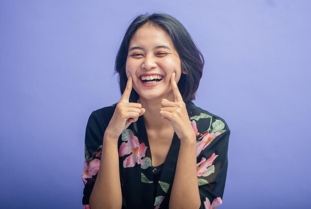Junge asiatische Frau mit fröhlicher Geste, die auf die Wange zeigt, isoliert auf violettem Hintergrund