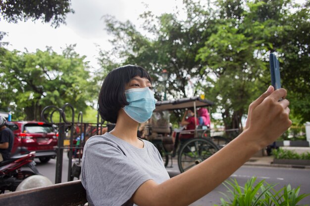 Junge asiatische Frau mit Gesichtsmaske mit Handy im öffentlichen Raum Corona-Virus-Konzept