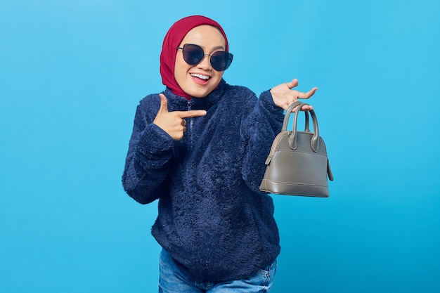 Junge asiatische Frau mit Jacke und lächelnder Sonnenbrille, die mit dem Finger auf die Tasche auf blauem Hintergrund zeigt