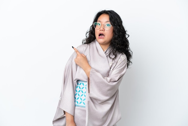 Junge asiatische Frau mit Kimono isoliert auf weißem Hintergrund überrascht und zeigt Seite