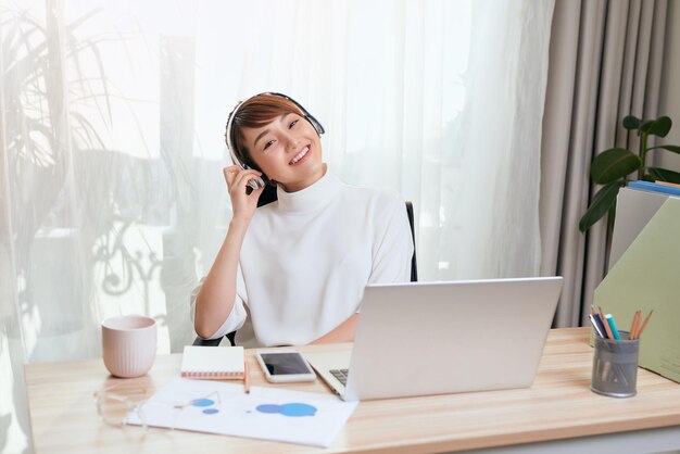 Junge asiatische Frau mit Kopfhörern, die zu Hause am Computer arbeitet