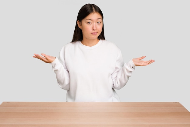 Junge asiatische Frau sitzt auf einem isolierten Tisch, zweifelt und zuckt mit den Schultern.
