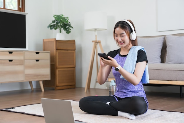 Junge asiatische Frau surft mit ihrem Smartphone im Internet und hört Musik, nachdem sie zu Hause Sport gemacht hat