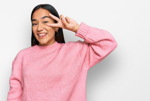 Junge asiatische Frau trägt einen lässigen Winterpullover und macht ein Friedenssymbol mit den Fingern über dem Gesicht, lächelt fröhlich und zeigt den Sieg