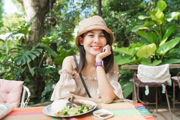 Junge asiatische Frau trägt Hut, lächelt und isst Mittagessen mit Fischfleisch im tropischen Garten