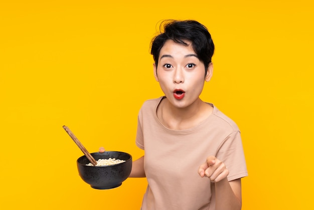 Junge asiatische Frau überrascht und zeigt nach vorne, während sie eine Schüssel Nudeln mit Stäbchen hält
