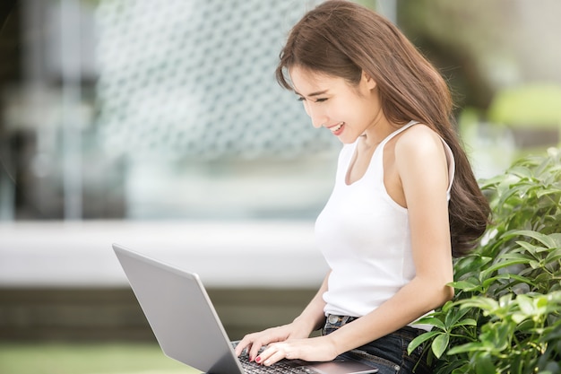 Junge asiatische freiberuflich tätige Geschäftsfrau, die an ihrem Laptop beim Sitzen im Stadtpark arbeitet