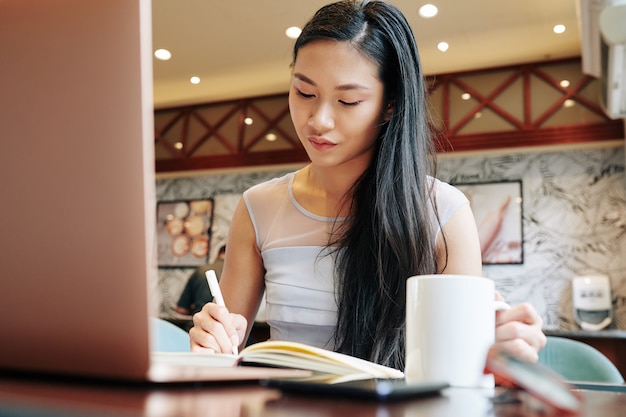 Junge asiatische Geschäftsfrau, die Planer beim Trinken des Morgenkaffees im Café füllt