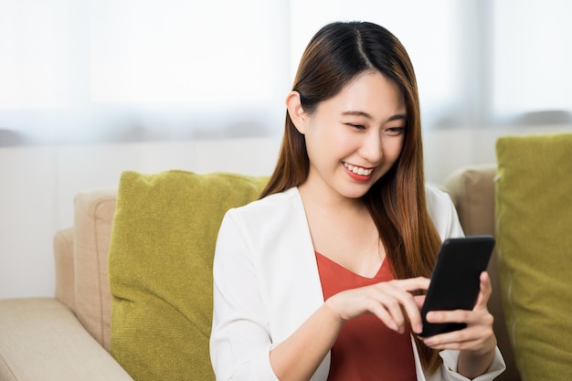 Junge asiatische Geschäftsfrau, die zu Hause mit Smartphone auf dem Sofa im Wohnzimmer sitzt und lächelt.