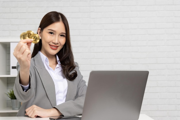 Junge asiatische Geschäftsfrau im Anzug sitzt und hält die goldene Bitcoin BTC-Kryptowährung in der Hand