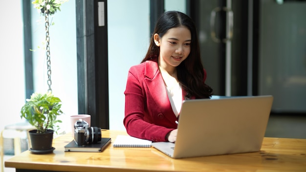 Junge asiatische Geschäftsfrau im eleganten roten Anzug sitzt im Büro und tippt und arbeitet an Laptop-Computern