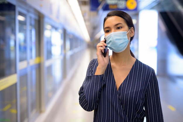 Junge asiatische Geschäftsfrau mit Maske, die am Telefon am U-Bahn-Bahnhof spricht