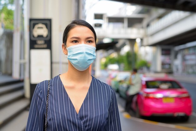 Junge asiatische Geschäftsfrau mit Maske, die an der Taxistation in den Stadtstraßen wartet