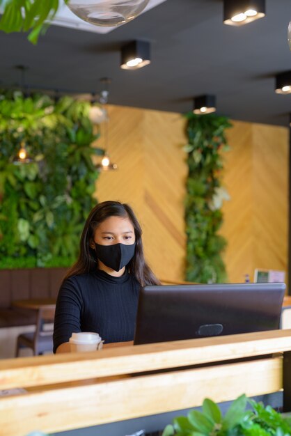 Junge asiatische Geschäftsfrau mit Maske, die Laptop innerhalb des Kaffeehauses verwendet