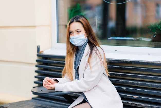 Junge asiatische Geschäftsfrau mit Maske, die Telefon benutzt und mit Abstand außerhalb des Coffeeshops sitzt