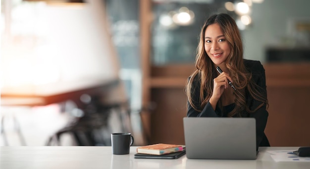 Junge asiatische Geschäftsfrau schön charmant lächelnd mit Stift und Arbeiten am Laptop-Computer im Büro Lächelnd und Blick in die Kamera