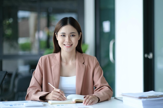 Junge asiatische Geschäftsfrau sitzt an ihrem Schreibtisch und macht sich Notizen mit Laptop im Büro