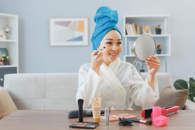 Junge asiatische glückliche frau mit handtuch auf dem kopf, die am schminktisch zu hause innen sitzt und puder oder rouge aufträgt, der spiegel hält, der morgens make-up-routine macht