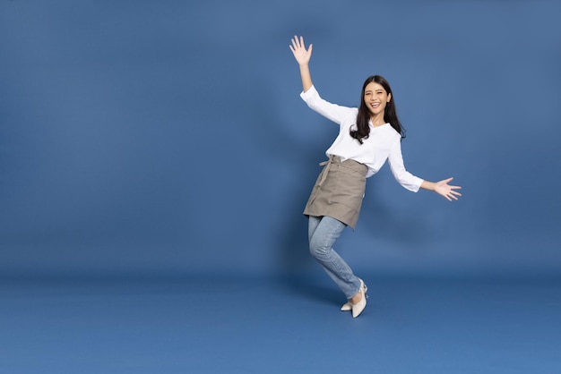 Junge asiatische Hausfrau mit Küchenschürze, die isoliert auf blauem Hintergrund kocht und tanzt