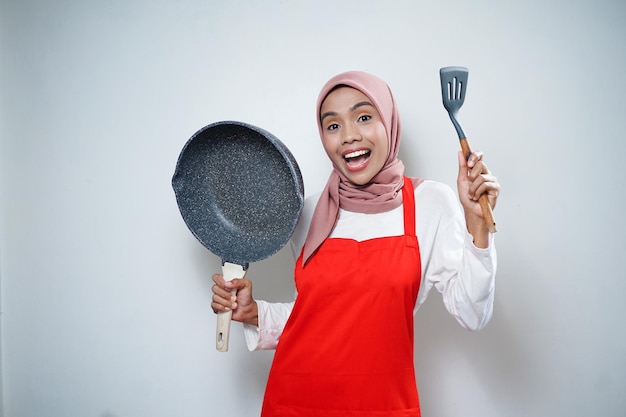 Junge asiatische Hausfrau mit Küchenschürze, die Pfanne und Pfannenwender kocht und hält