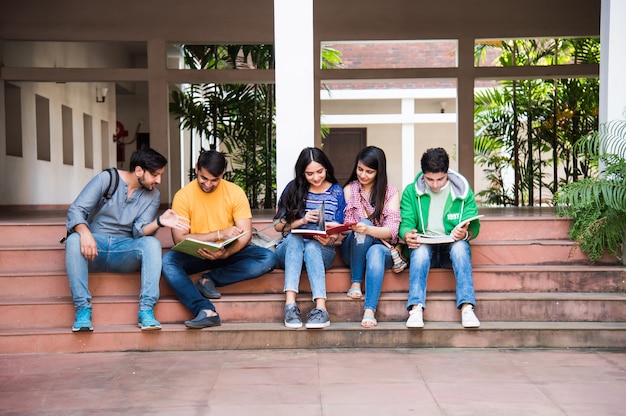 Junge asiatische indische College-Studenten, die Bücher lesen, am Laptop lernen, sich auf die Prüfung vorbereiten oder an Gruppenprojekten arbeiten, während sie auf Gras, Treppen oder Stufen des College-Campus sitzen sitting