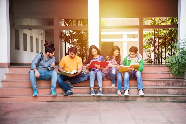 Junge asiatische indische College-Studenten, die Bücher lesen, am Laptop lernen, sich auf die Prüfung vorbereiten oder an Gruppenprojekten arbeiten, während sie auf Gras, Treppen oder Stufen des College-Campus sitzen sitting