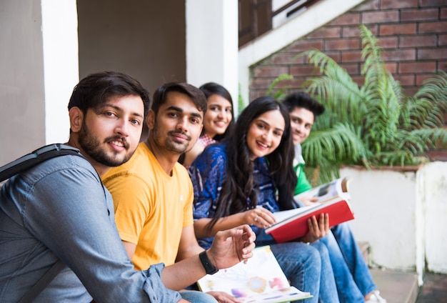 Junge asiatische indische college-studenten, die bücher lesen, am laptop lernen, sich auf die prüfung vorbereiten oder an gruppenprojekten arbeiten, während sie auf gras, treppen oder stufen des college-campus sitzen sitting