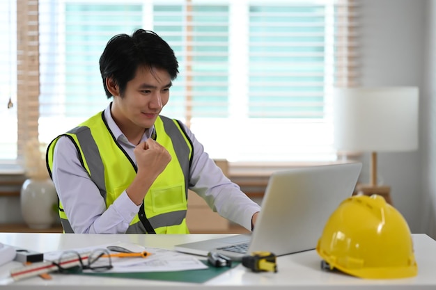 Junge asiatische Ingenieurarbeiterin in reflektierender Jacke, die auf Laptop-Computer schaut und Erfolg feiert