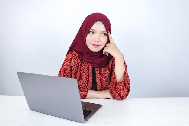 Junge asiatische Islamfrau lächelt mit denkender Geste und schaut auf die Leerstelle vor dem Laptop