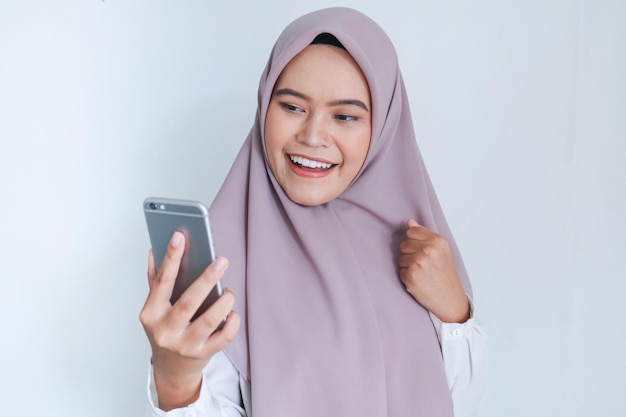 Junge asiatische Islamfrau mit Kopftuch feiert glücklich und aufgeregt das, was sie auf dem Smartphone sieht Indonesische Frau auf grauem Hintergrund