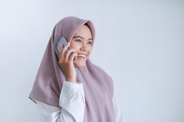 Junge asiatische Islamfrau mit Kopftuch ist glücklich und lächelt, wenn sie am Telefon spricht Indonesische Frau auf grauem Hintergrund