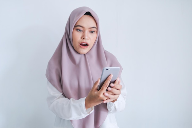 Junge asiatische Islamfrau mit Kopftuch ist schockiert und wow im Smartphone Indonesische Frau auf grauem Hintergrund