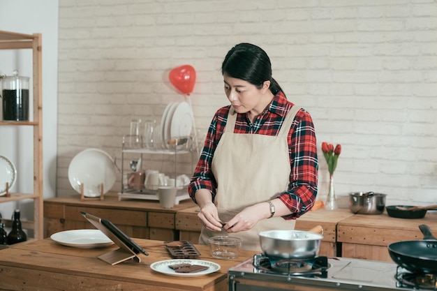 Junge asiatische Köchin, gekleidet in Schürze, macht handgemachte Pralinen in der Küche in einer gemütlichen Wohnung. elegantes mädchen im hemd, das rezeptvideo auf digitalem online-tablet am valentinstag anschaut.