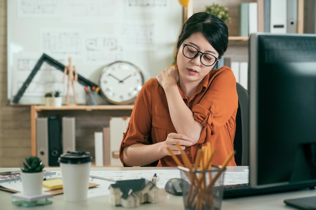 Junge asiatische koreanische mitarbeiterin leidet unter schulterschmerzkonzept-bürosyndrom. unglückliche, müde Architektin Arbeitermassage, die den Hals verletzt, sitzt am Schreibtisch und starrt lange Zeit auf den Computerbildschirm