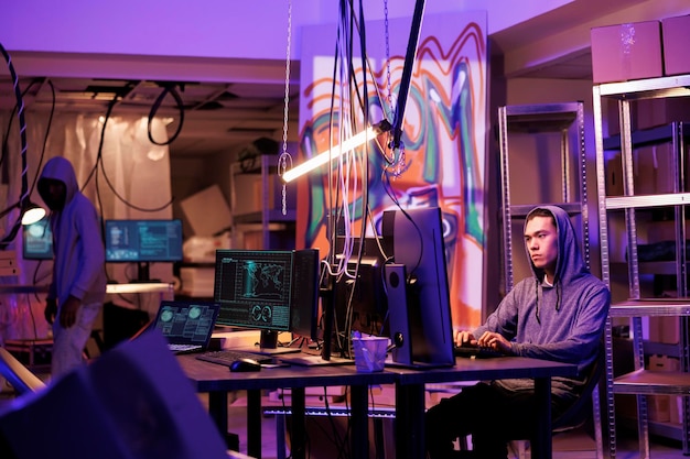 Junge asiatische Kriminelle benutzen Computer, um das System zu hacken und planen einen Cyberangriff, um Daten zu stehlen Hacker in der Kapuze entwickelt illegale bösartige Software in einem verlassenen Lagerhaus in der Nacht