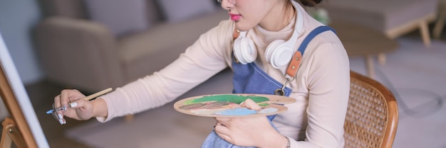 Junge asiatische Künstlerin trägt Kopfhörer am Hals und malt mit dem Pinsel Kunstwerke auf Leinwand