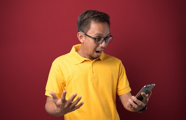 Junge asiatische Mann mit Brille mit Smartphone auf rotem Hintergrund schockiert mit einem Überraschungsgesicht.