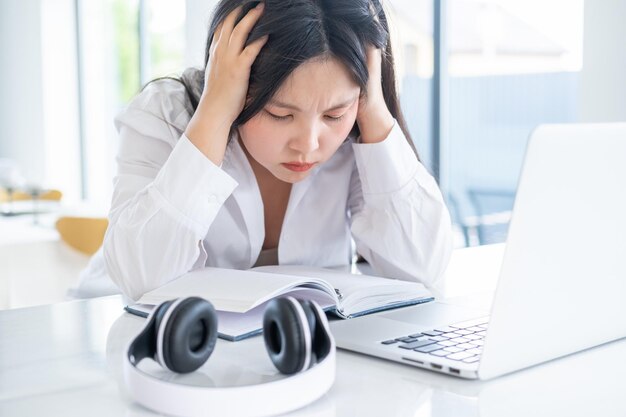 Junge asiatische müde Geschäftsfrau mit langen Haaren, die am Laptop im hellen Heimbüro arbeitet