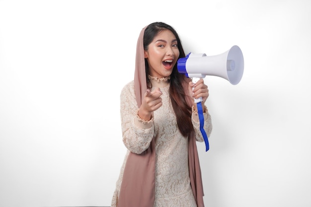 Junge asiatische muslimische Frau mit Kopftuch, Schleier-Hijab, spricht durch ein Megafon, während sie auf die Kamera zeigt, isoliert auf weißem Hintergrund Studio Ramadan und Eid Mubarak Konzept