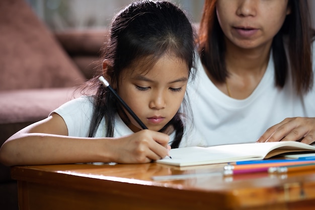 Junge asiatische Mutter hilft ihrer Tochter, ihre Hausaufgaben zu Hause zu machen. Bildung von zu Hause Konzept.