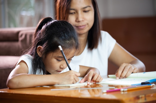 Junge asiatische Mutter hilft ihrer Tochter, ihre Hausaufgaben zu Hause zu machen. Bildung von zu Hause Konzept.