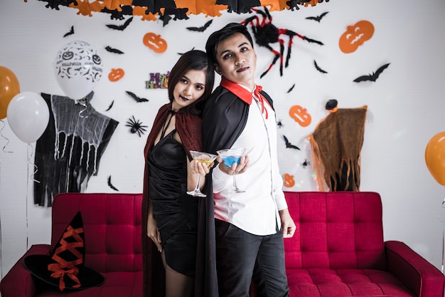 Junge asiatische Paare in Kostümhexe und Dracula mit feiern Halloween-Party und trinken Wein zusammen beim Halloween-Festival im Zimmer zu Hause. Konzeptpaare feiern Halloween zusammen zu Hause.