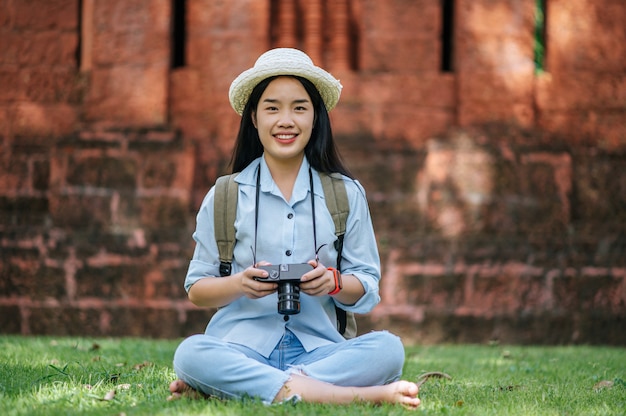 Junge asiatische Rucksacktouristin mit Hut, die lächelt, während sie eine Reise in die historische Stätte hat, sitzt auf Gras zum Entspannen und benutzt die Kamera, um ein Foto mit glücklichen zu machen