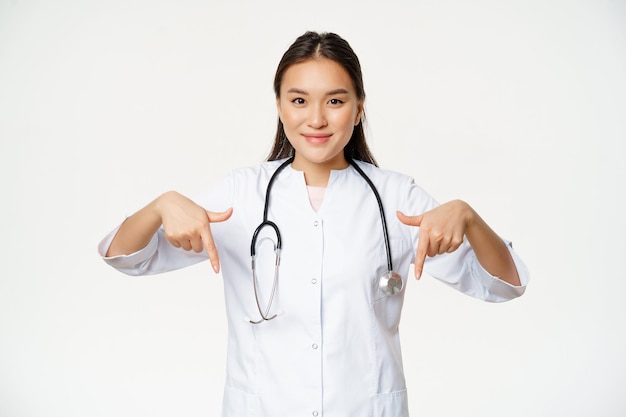 Junge asiatische Ärztin, die mit den Fingern nach unten zeigt und lächelt, Stethoskop und medizinische Uniform trägt und auf weißem Hintergrund steht
